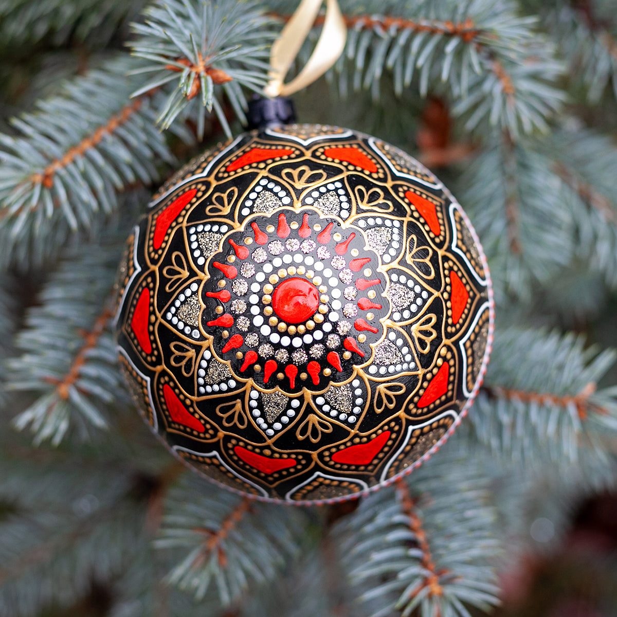 Close-up view of a red Christmas ornament with hand-painted mandala design. Blue fir tree on the background.