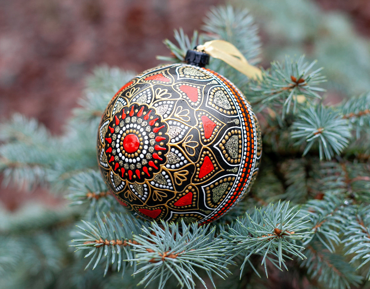 Close-up view of a red Christmas ornament with hand-painted mandala design. Blue fir tree on the background.