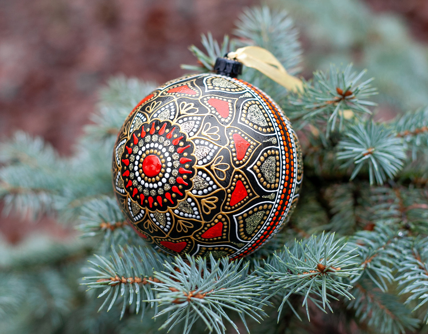 Close-up view of a red Christmas ornament with hand-painted mandala design. Blue fir tree on the background.