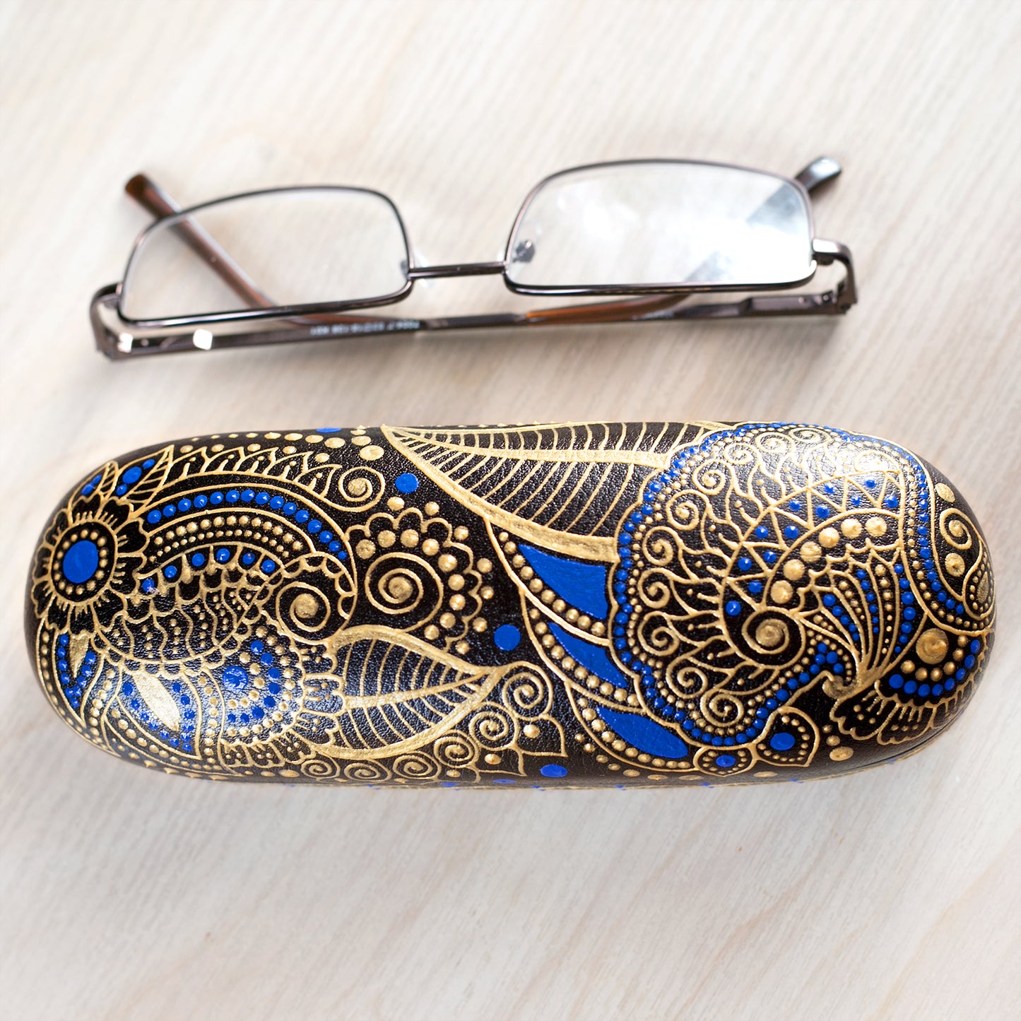 Hardshell case for glasses, hand-painted with blue and gold mehndi design. View from the top, reading glasses are on the side to show dimensions of the case.