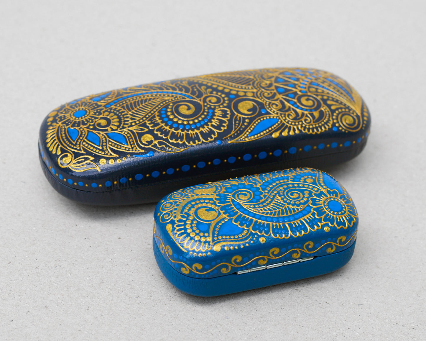 Hardshell case for glasses, hand-painted with blue and gold mehndi design. Set of eyeglasses case and earphones case on a gray background