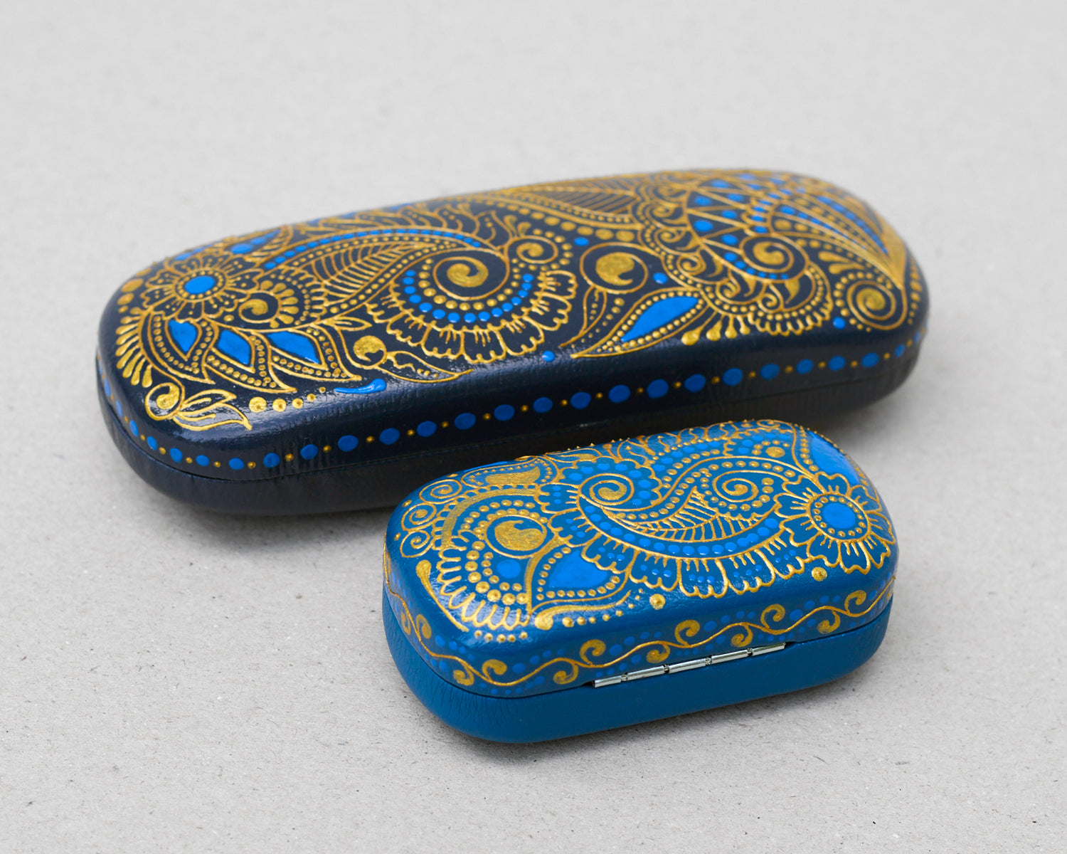 Hardshell case for glasses, hand-painted with blue and gold mehndi design. Set of eyeglasses case and earphones case on a gray background