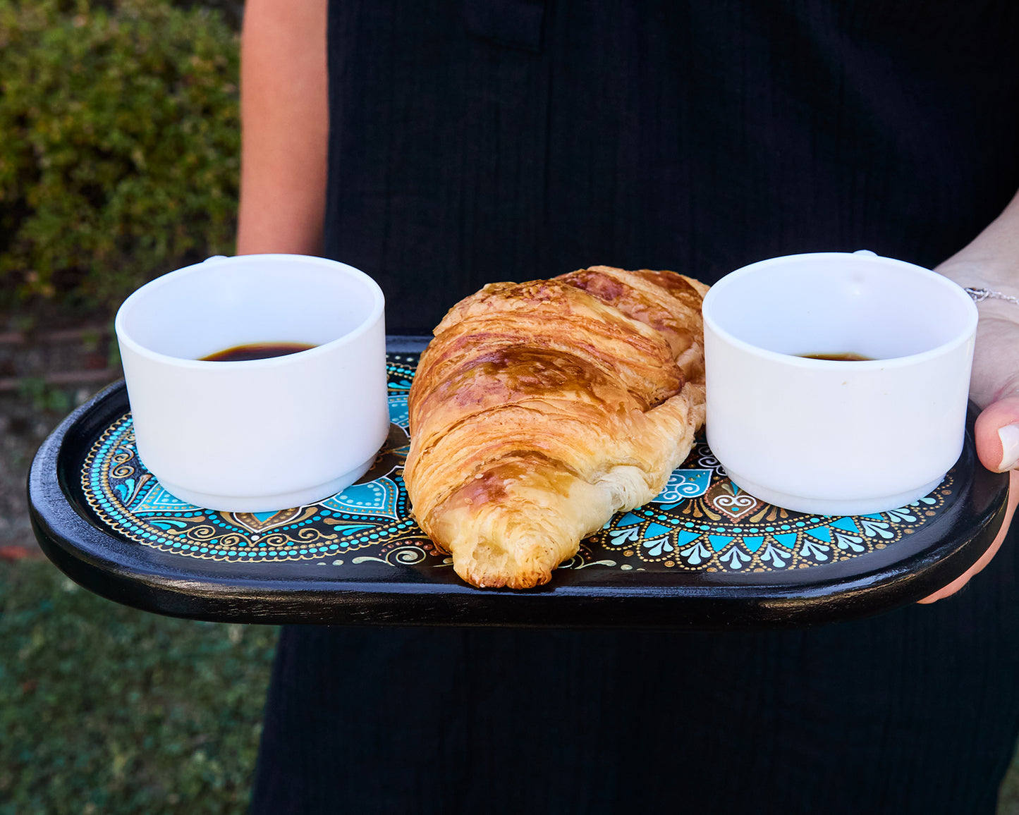 Woman holds serving tray in moroccan style. Two coffee cups and a croissant on it.