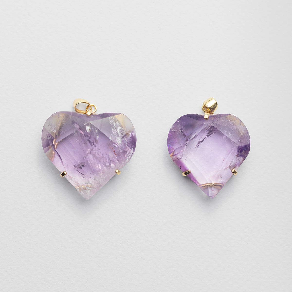 Two heart-shaped amethyst pendants with gold bail loops, displayed side-by-side on a light gray background.