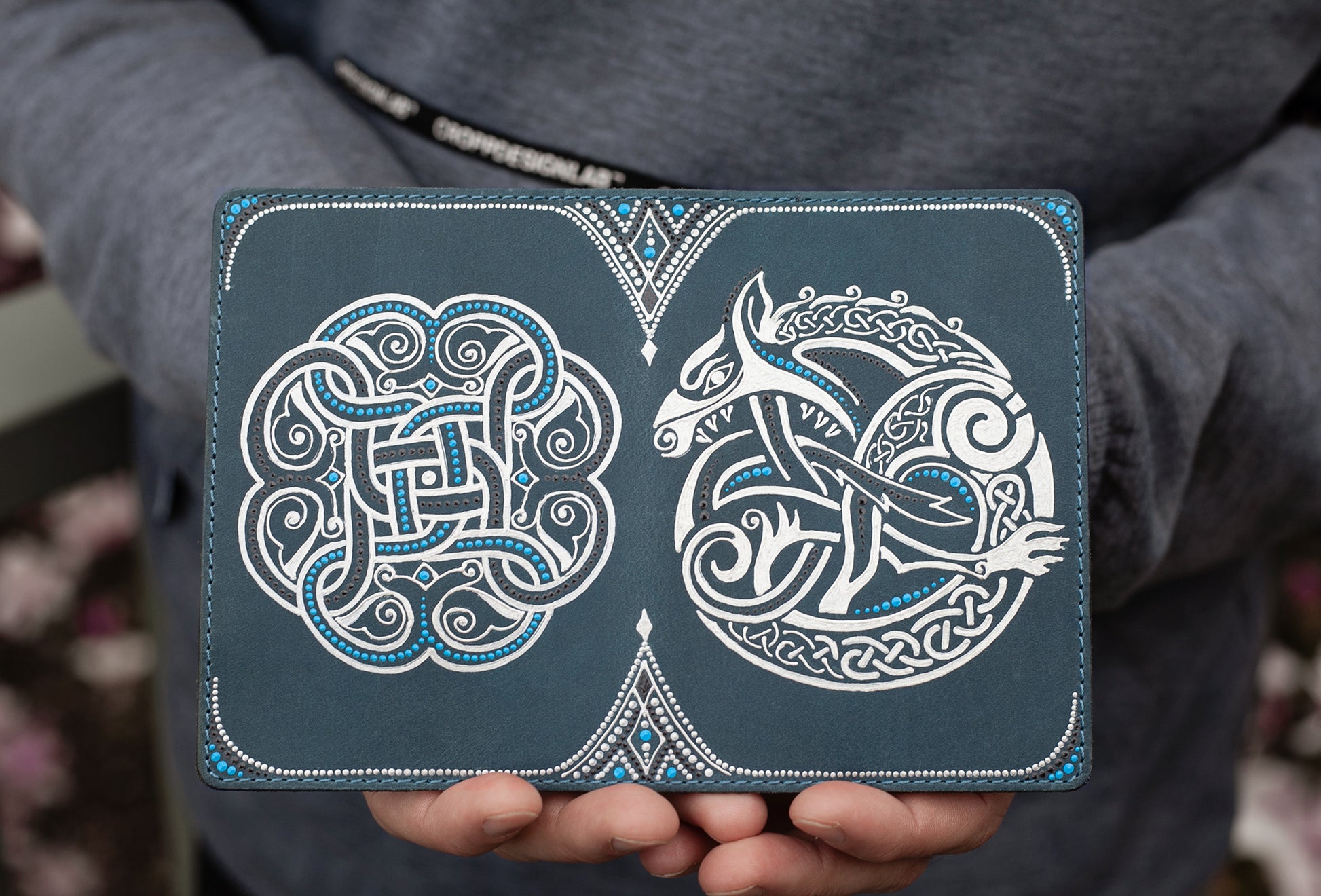 Man holding open passport cover. The color is gray and silver, with hand-painted celtic picture of Fenrir. Close-up view.