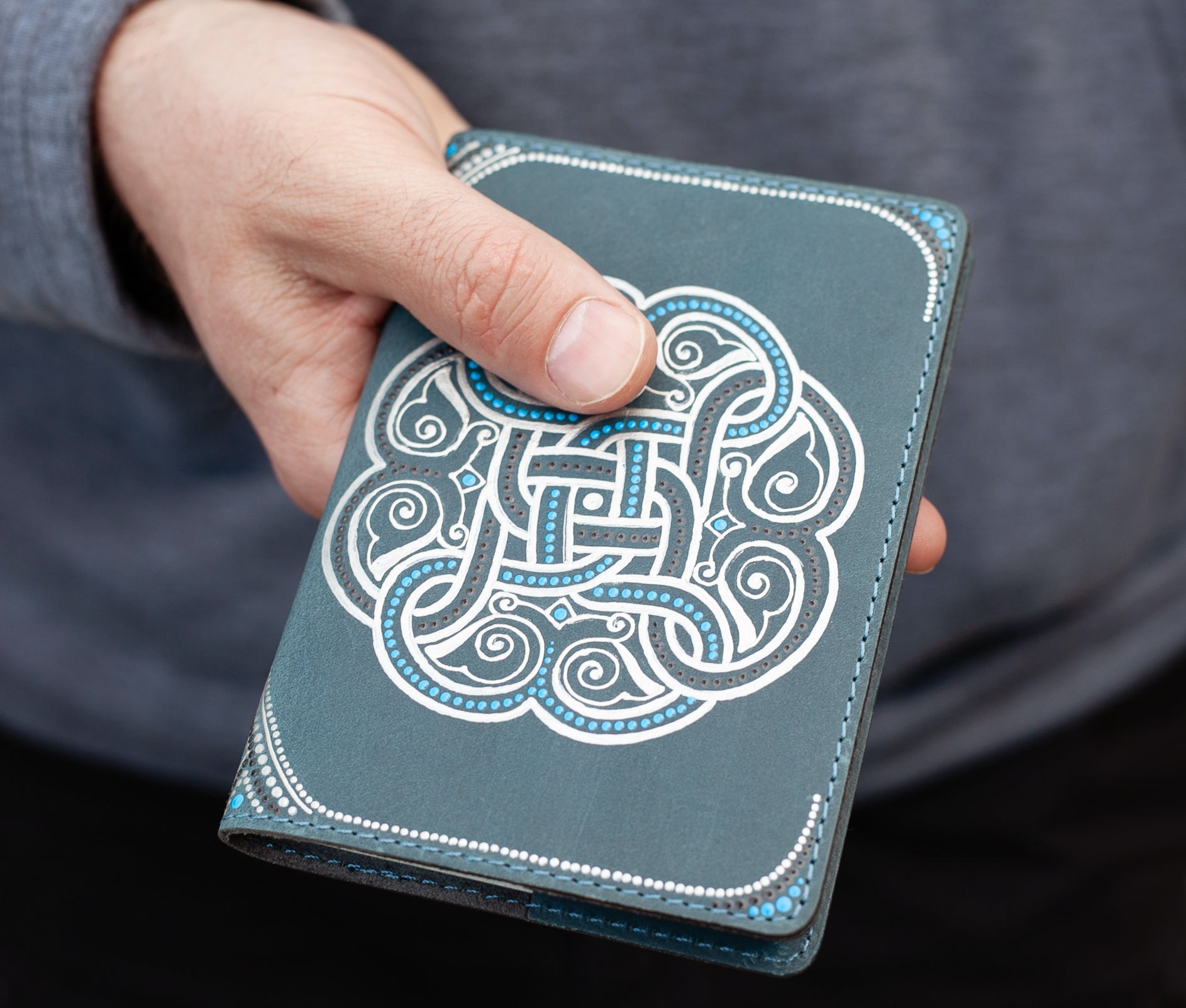 Man holding in hand leather passport cover. The color is gray and silver, with hand-painted celtic knot. Close-up view.