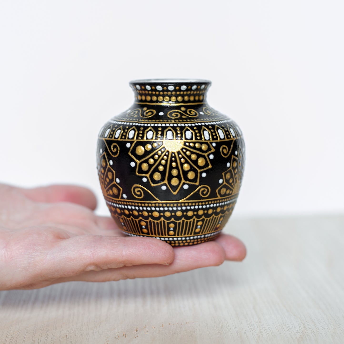 A small, hand-painted ceramic bud vase with greek style gold patterns, held in the palm of a hand.