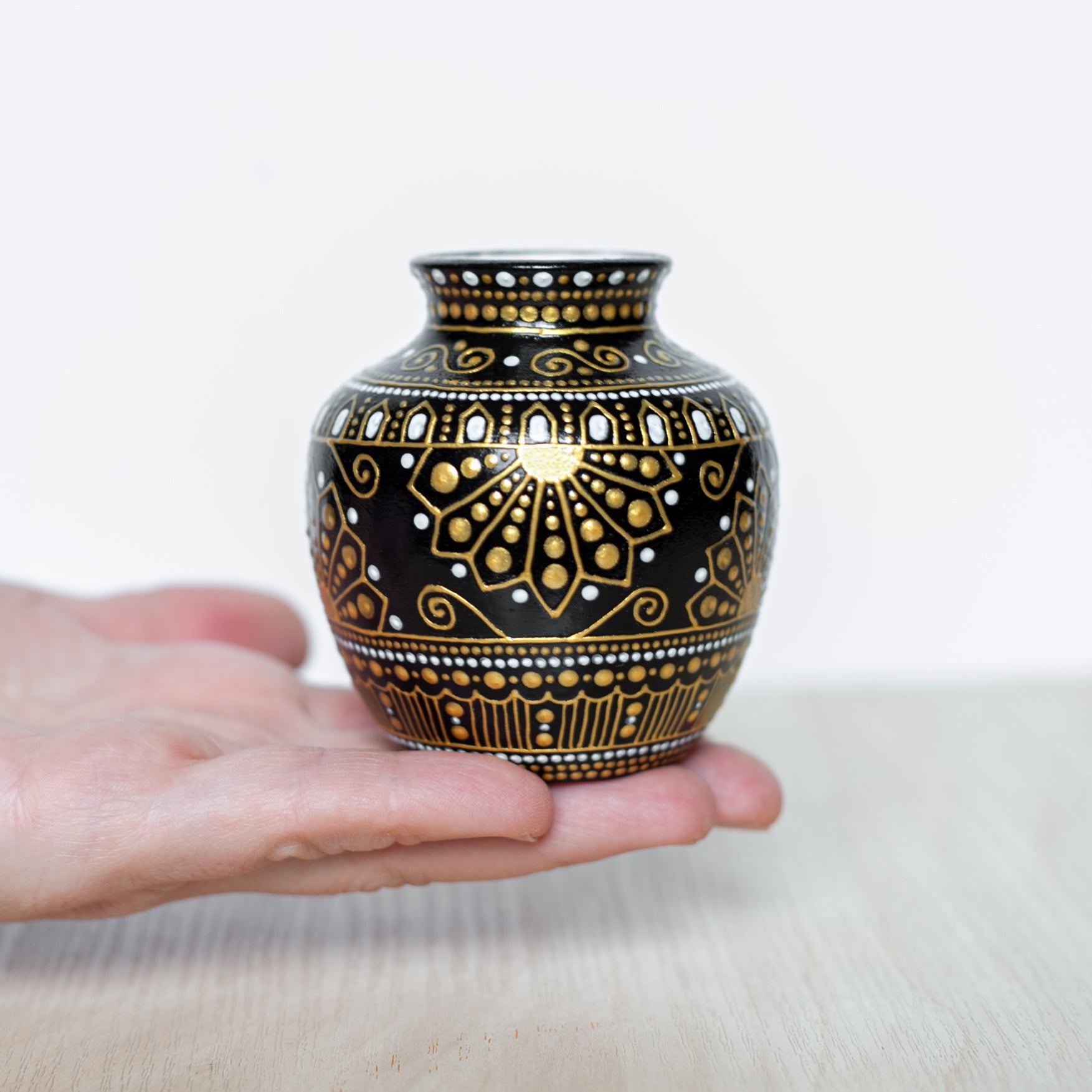 A small, hand-painted ceramic bud vase with greek style gold patterns, held in the palm of a hand.
