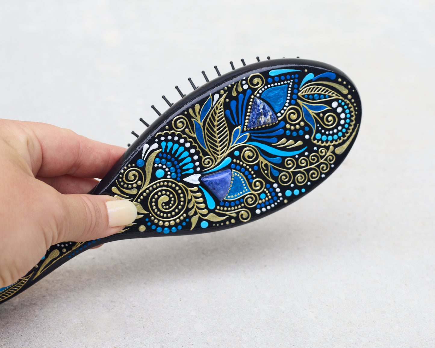 Hand holding a hand-painted wooden paddle hairbrush with blue floral design.