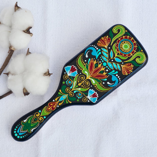 A colorful hand-painted wooden paddle hairbrush with folk floral patterns in red, green, blue, and gold on a black background. View from the top on a white linen cloth