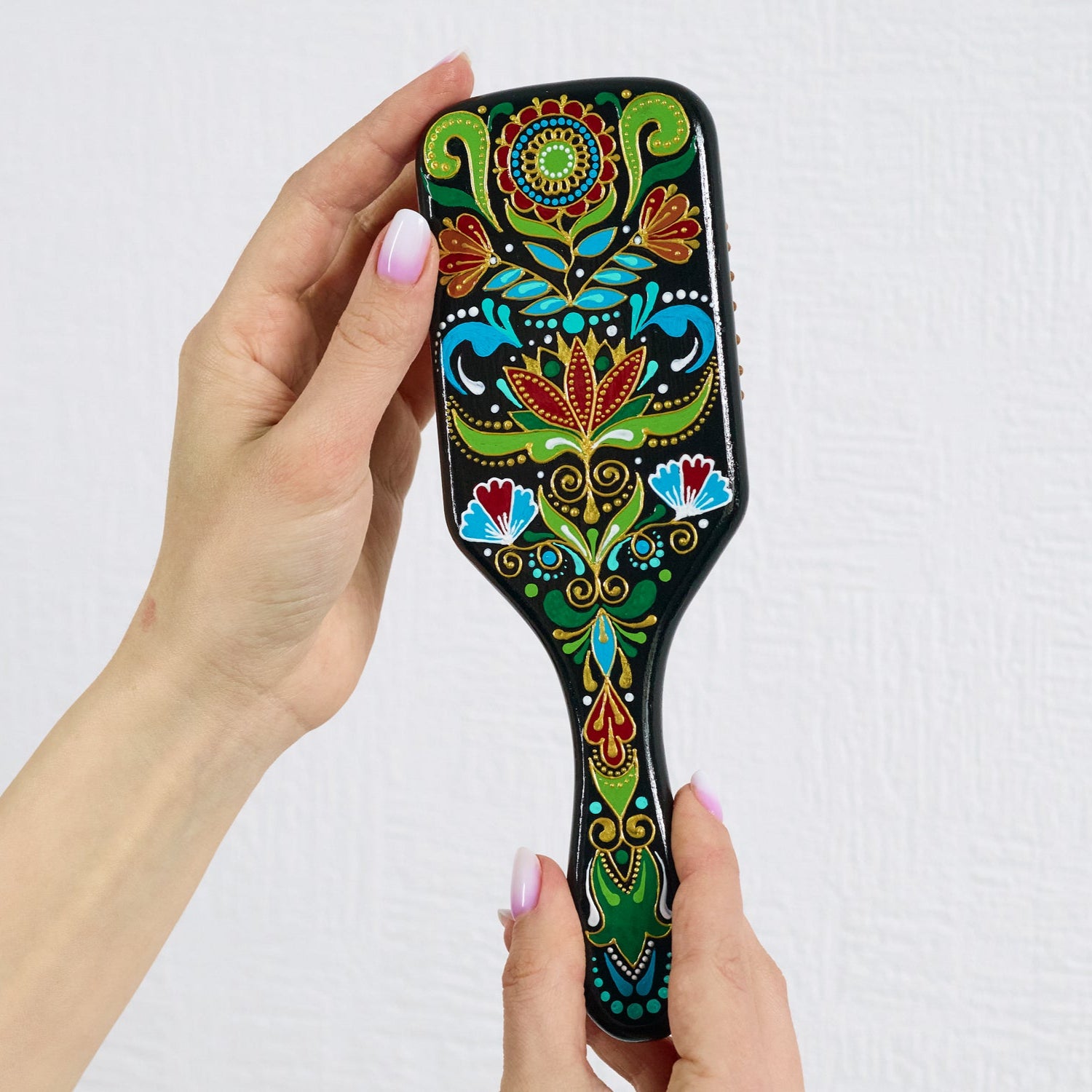 Hand holding a colorful hand-painted wooden paddle hairbrush with folk floral patterns in red, green, blue, and gold on a black background.
