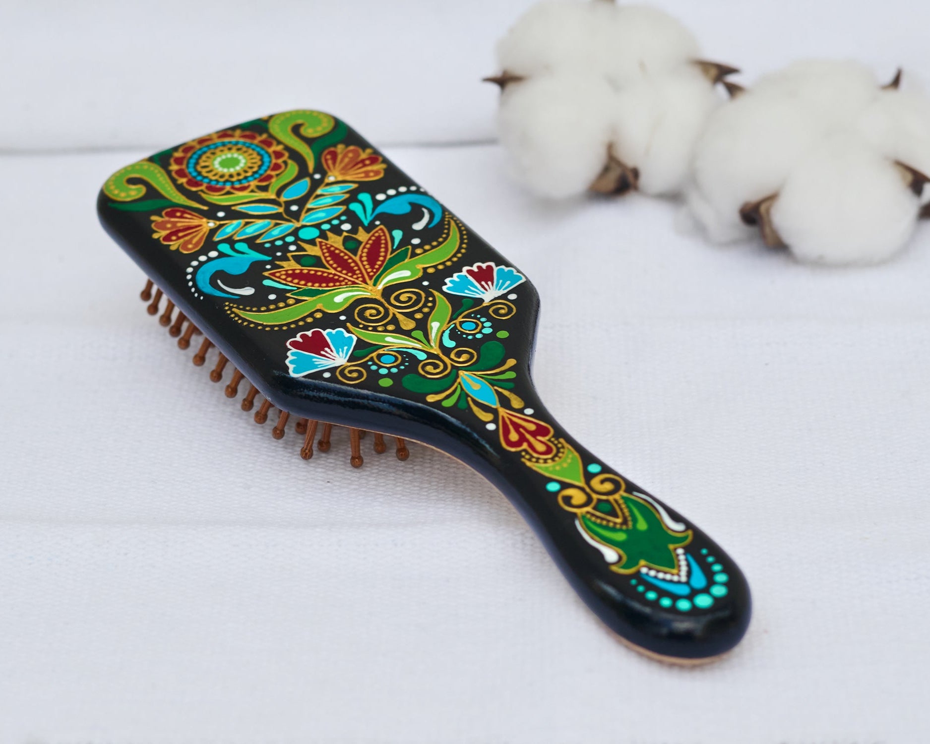 A colorful hand-painted wooden paddle hairbrush with folk floral patterns in red, green, blue, and gold on a black background. Close-up view, lying on a white linen cloth