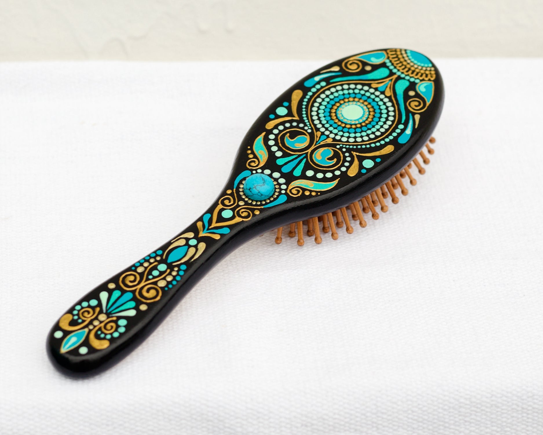 Hand-painted wooden paddle hairbrush with turkish design in turquoise blue and gold on a black background. View from the top on a white linen cloth