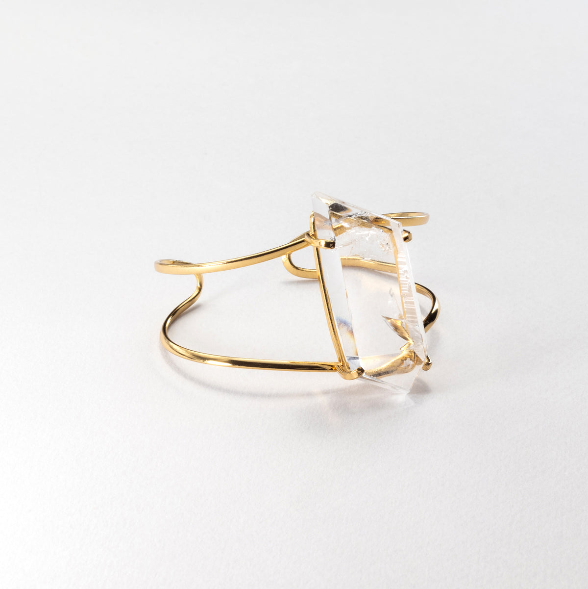 A gold-plated metal cuff ring featuring a large, clear, freeform faceted gemstone, photographed on a clean white background.