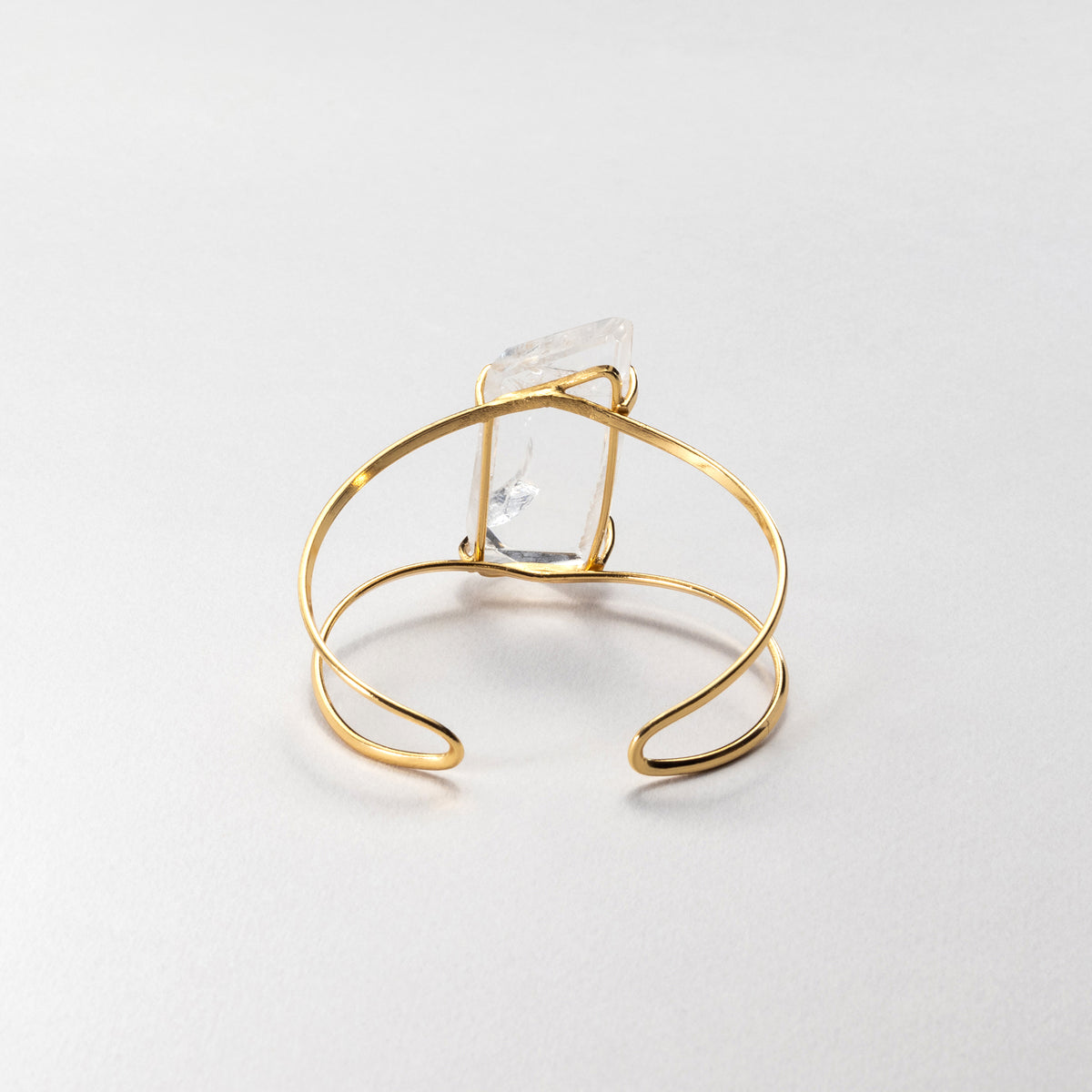 A gold wire wrap cuff bracelet featuring a clear, faceted rectangular crystal, photographed on a neutral light gray background.