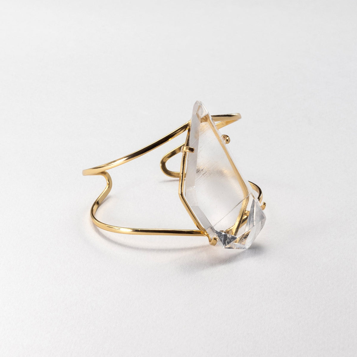 A gold-toned metal cuff bracelet featuring a large, faceted clear crystal gemstone, photographed on a clean white background.