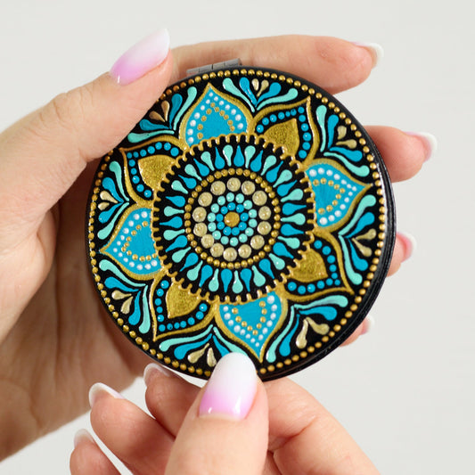 Woman's hands holding compact closed mirror showing its turquoise blue mandala hand-painted design. Close-up view