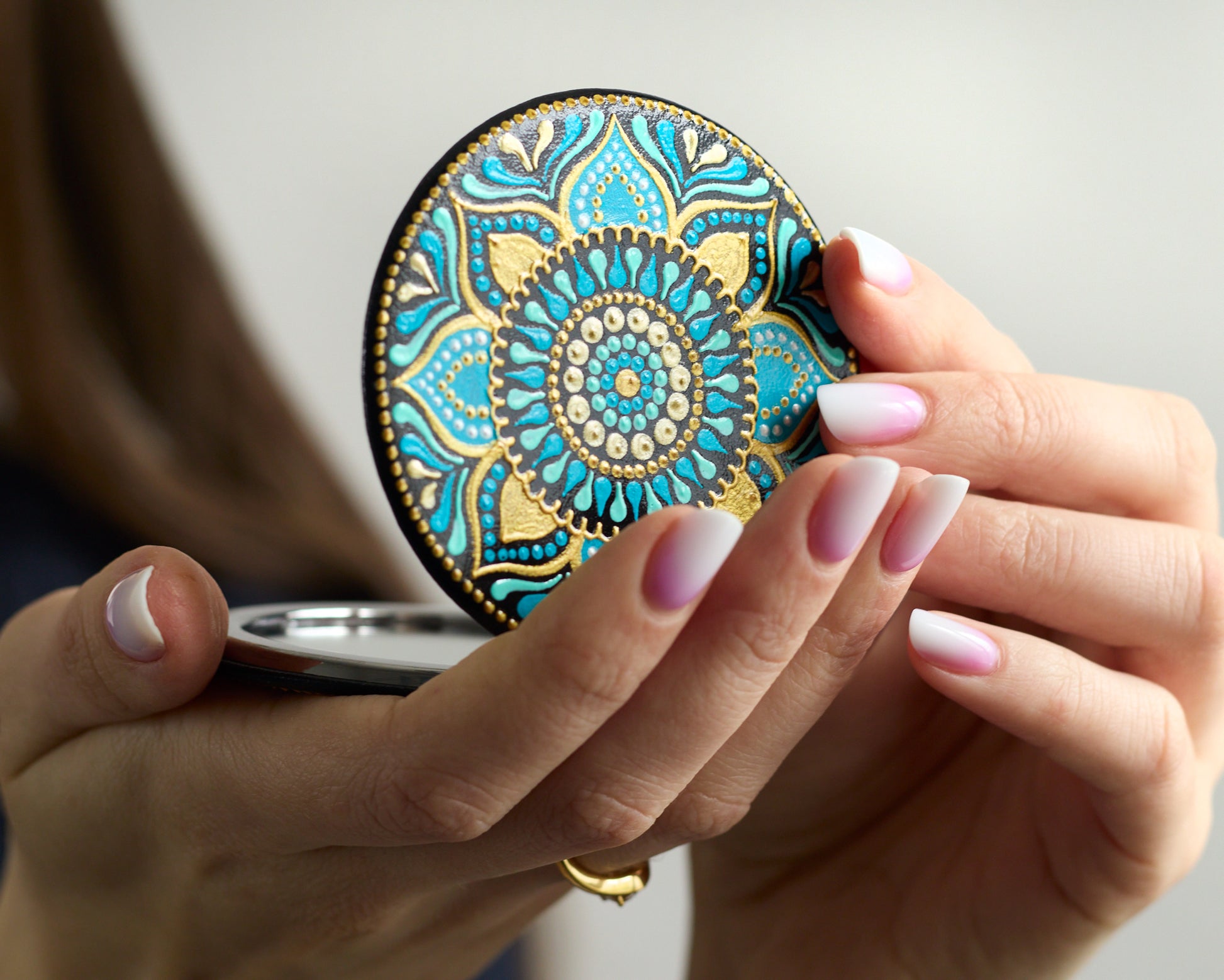 Woman holding in her hands opened compact mirror with turquoise blue mandala hand-painted design.