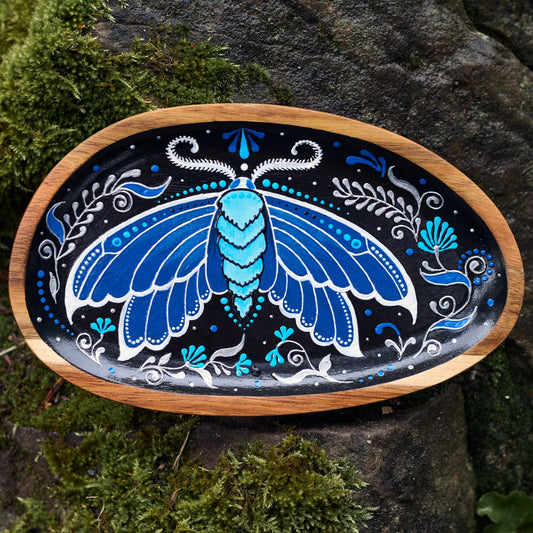 Top view of a small wooden plate with a hand-painted blue moth, surrounded by real moths on a natural textured background.