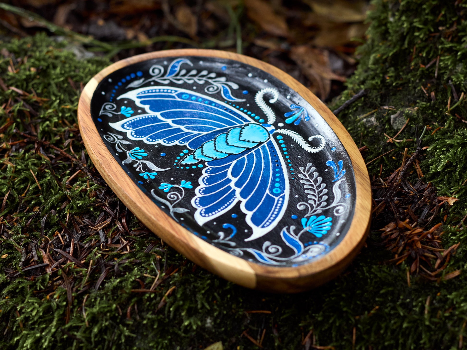 Side view of a small wooden plate with a painted blue moth, showing the thickness of the wood, placed on a natural background with real moths nearby