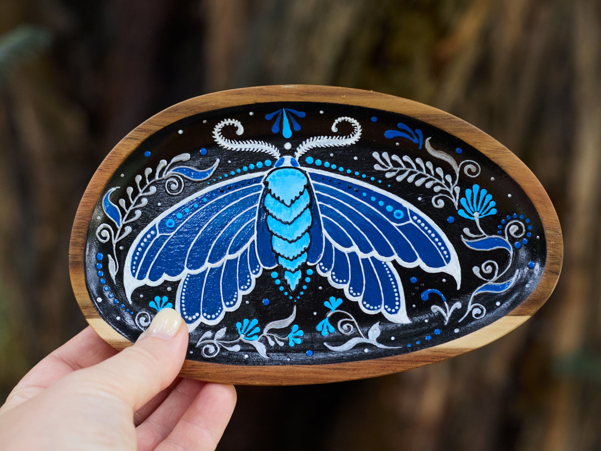 Close-up of a hand holding a wooden plate with blue moth illustration, highlighting handmade detail and natural, earthy vibe