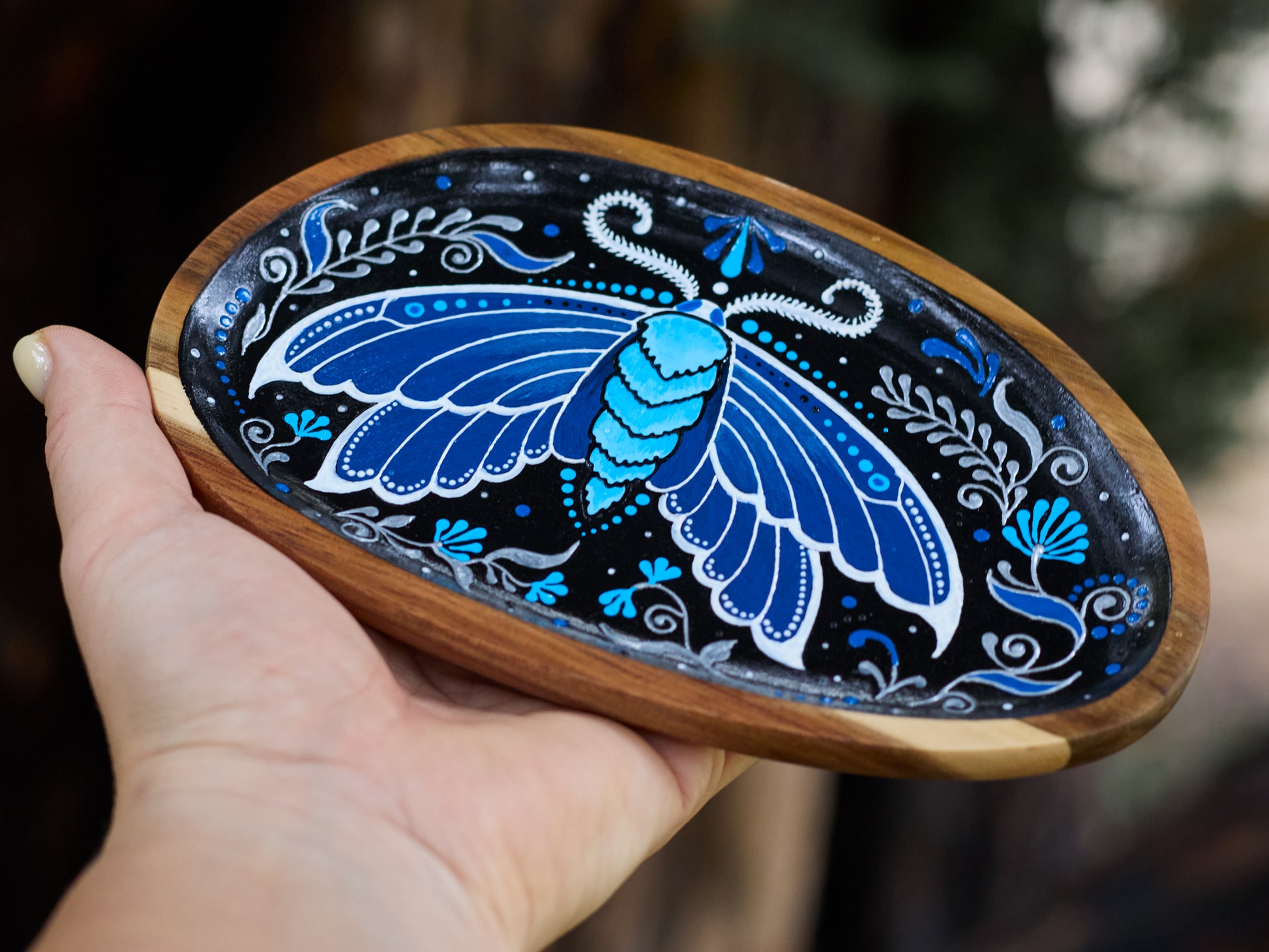 Close-up of a hand holding a wooden plate with blue moth illustration, highlighting handmade detail and natural, earthy vibe