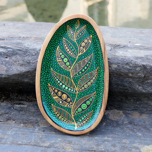Hand-painted wooden plate with a golden leaf design on a green dotted background created in the pointillism technique, placed on a stone surface outdoors.