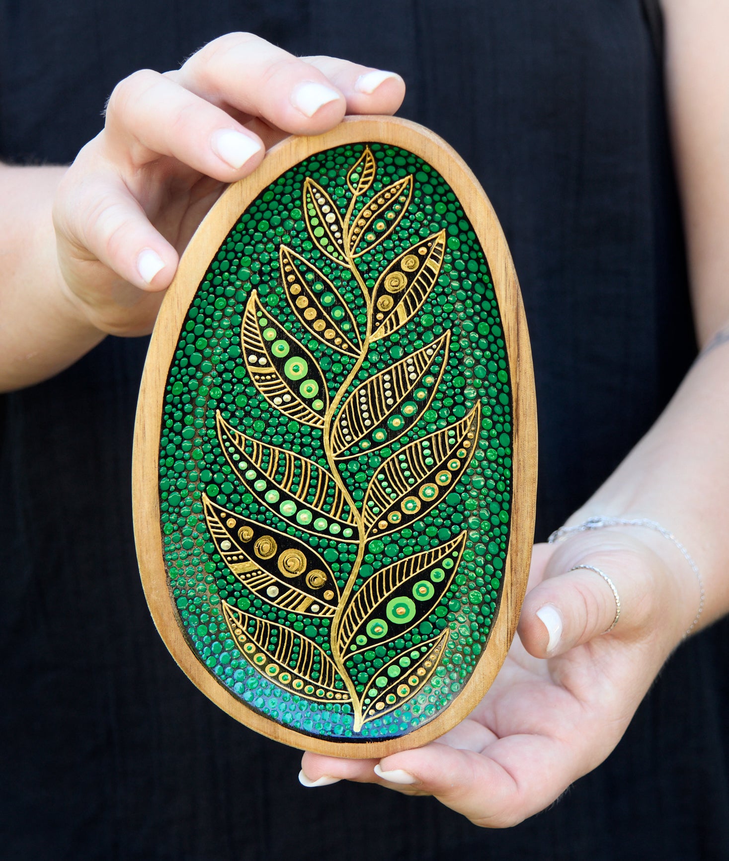 Hands holding small green wooden plate with a golden leaf design, black background.