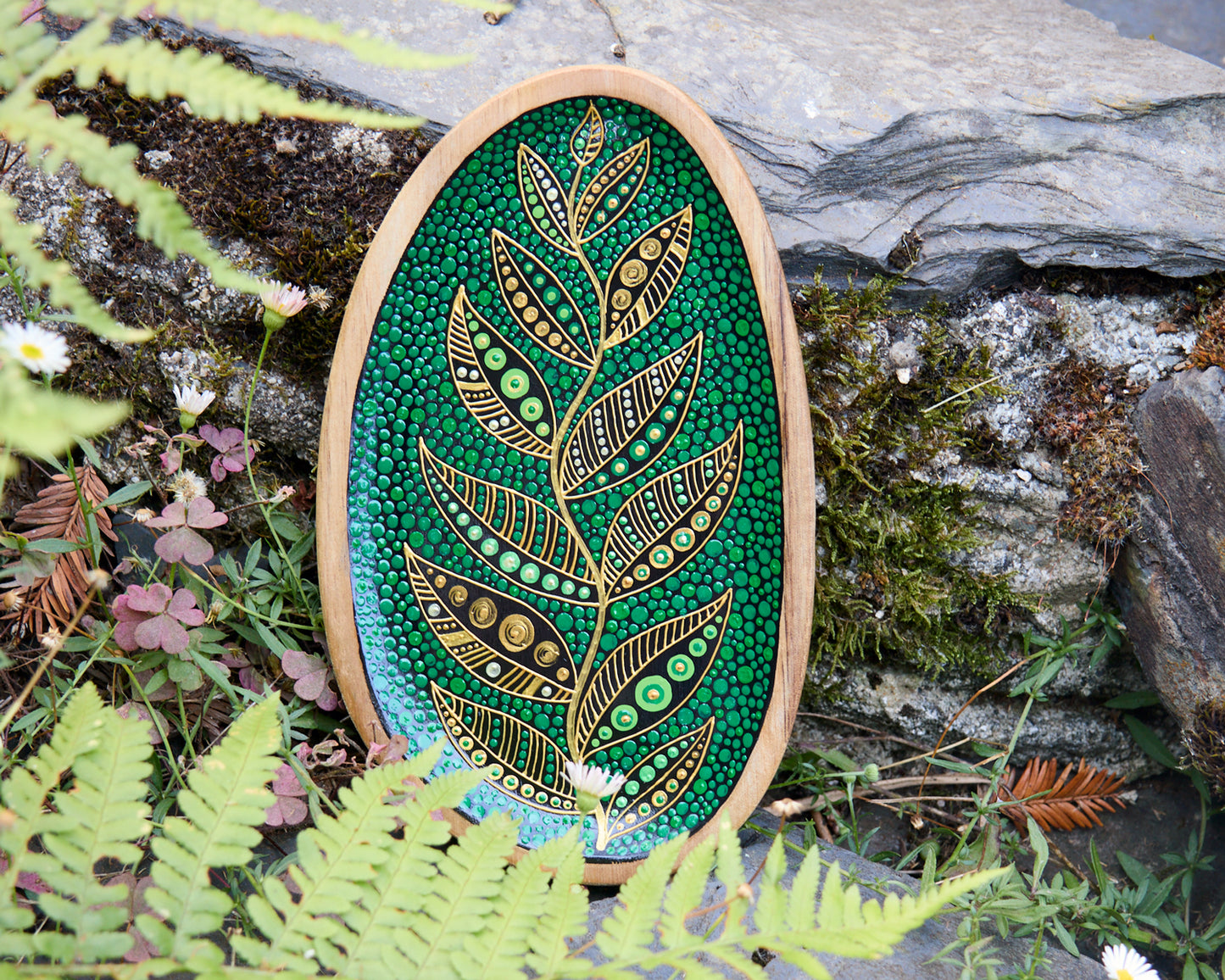 Hand-painted wooden plate with a golden leaf design on a green dotted background, placed on a stone surface outdoors.