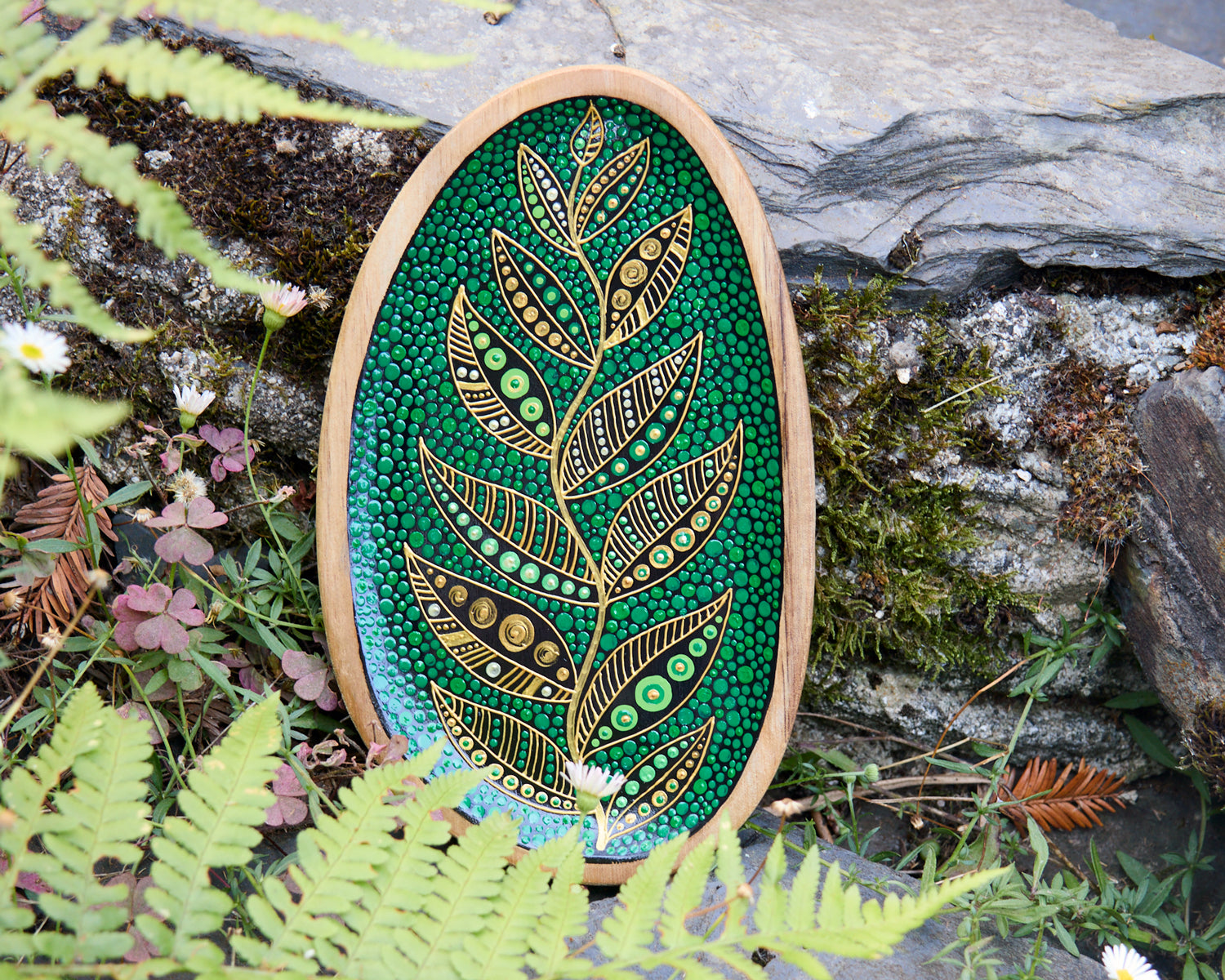 Hand-painted wooden plate with a golden leaf design on a green dotted background, placed on a stone surface outdoors.