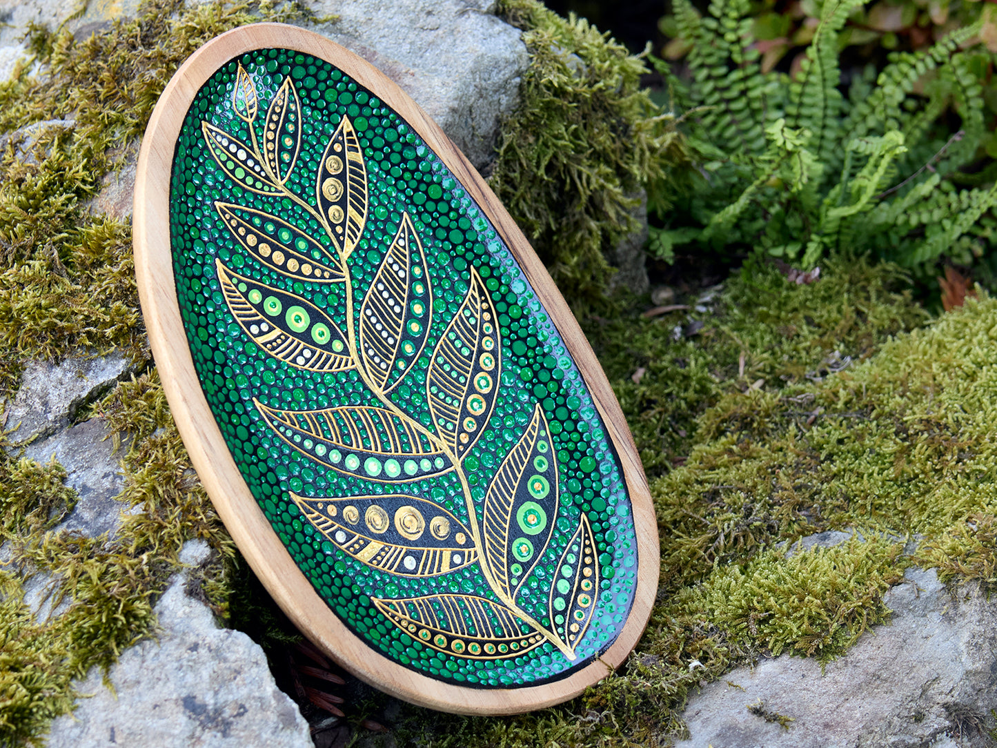Hand-painted wooden plate with a golden leaf design on a green dotted background, placed on a stone surface outdoors.