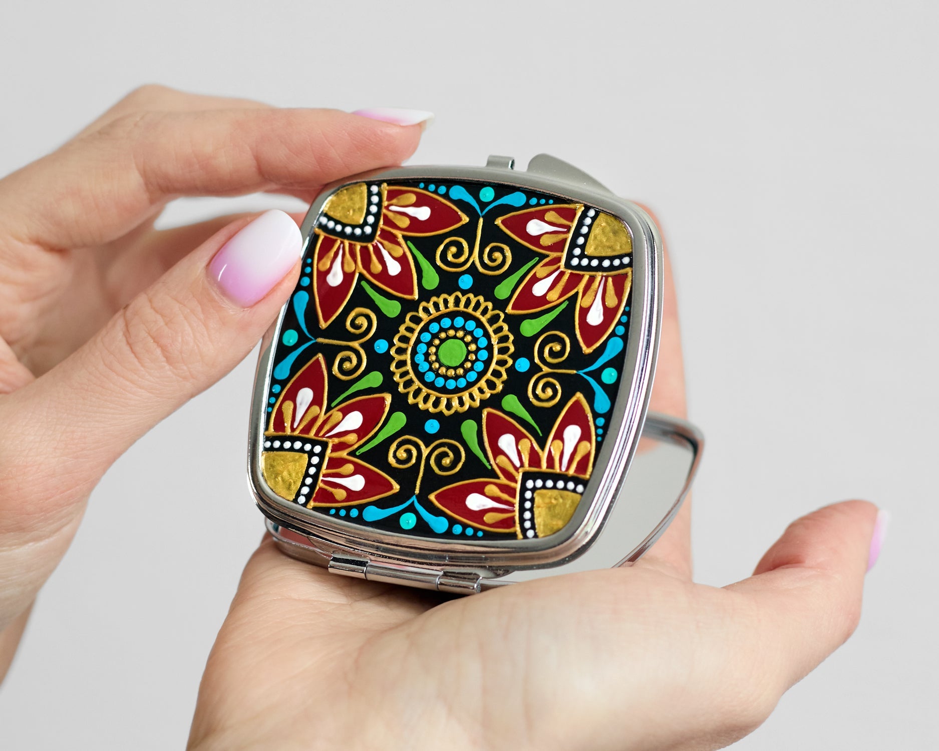 Hands holding an opened hand-painted compact mirror with folk floral design.