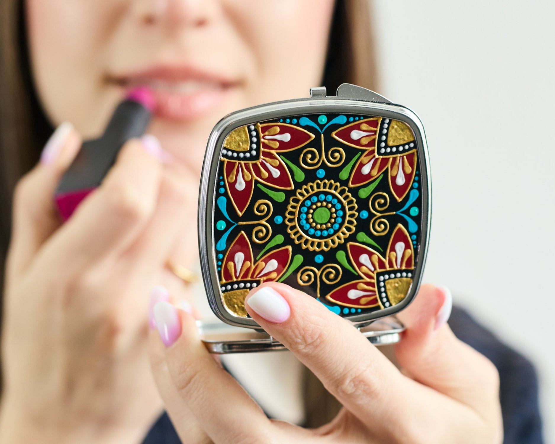 The woman holds her lipstick and looks into a compact mirror.