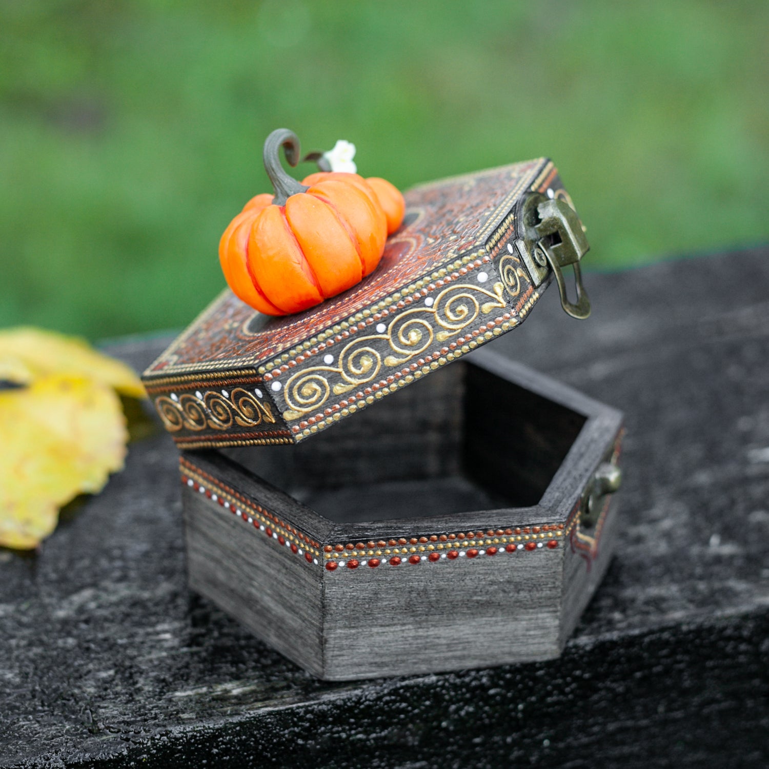 A hexagonal wooden box with hand-painted designs in gold and redis slightly open, revealing wooden interior. Two miniature pumpkins, made from polymer clay, attached to the top.  The box rests on a dark wooden surface with an autumn leaf beside it.