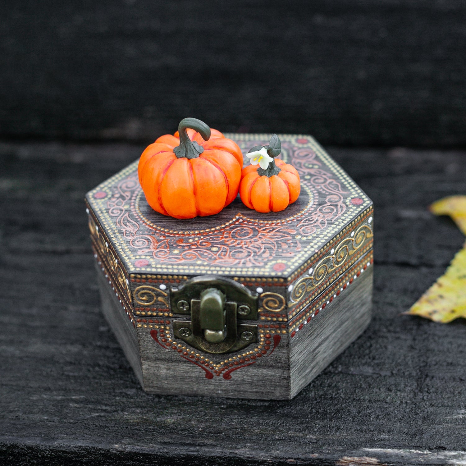 A hexagonal wooden box with hand-painted designs in gold and red sits on a mossy rock. Two miniature pumpkins, sculpted from polymer clay with remarkable detail, are attached to the top of the box. The background is a dark wood.