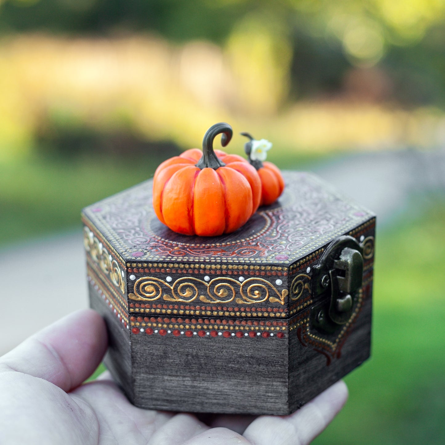 A hand holding a small, hexagonal wooden box decorated with hand-painted designs and two miniature pumpkins, sculpted from polymer clay.