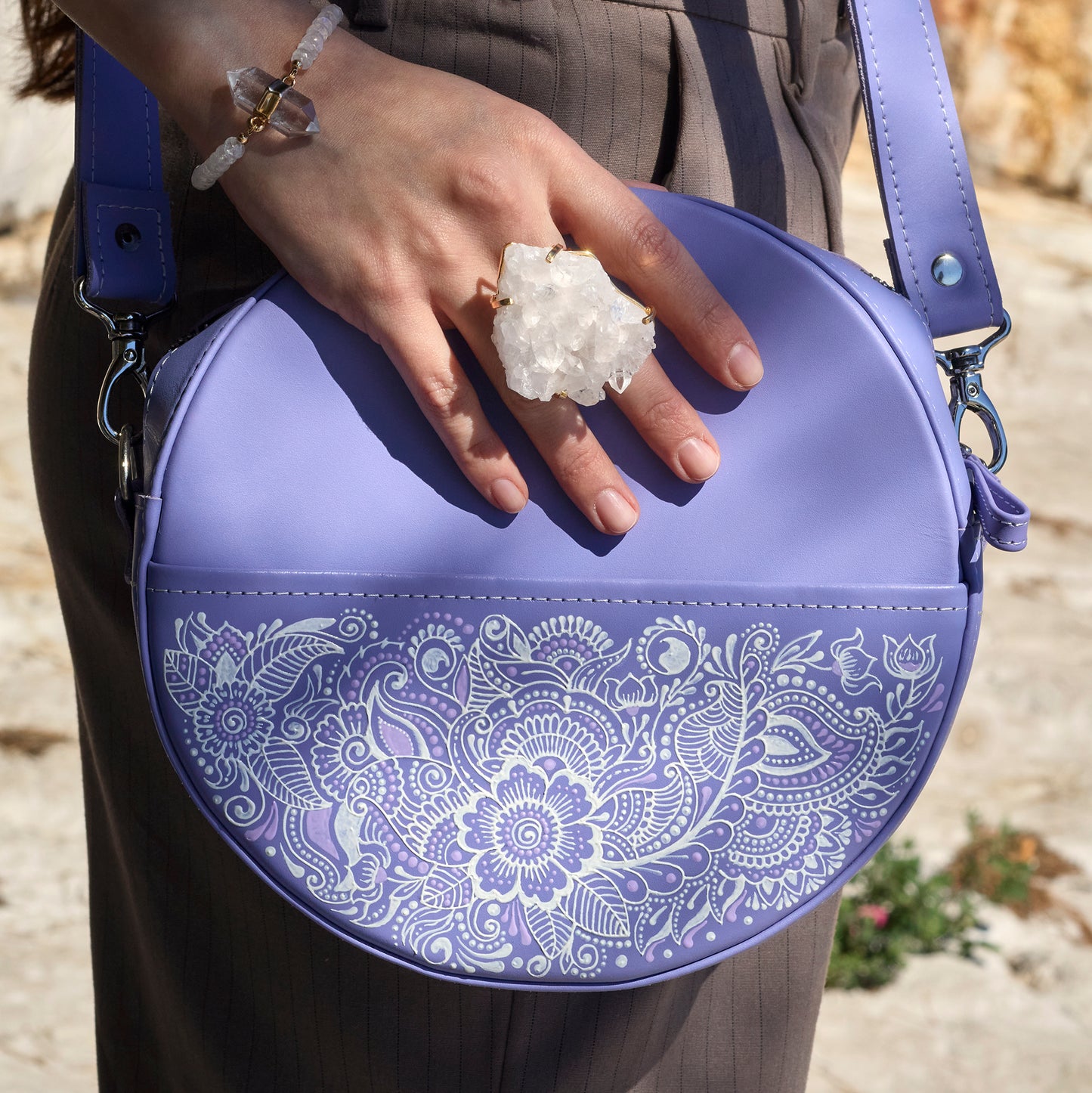 Person holding a purple round bag with white floral patterns, wearing a ring.
