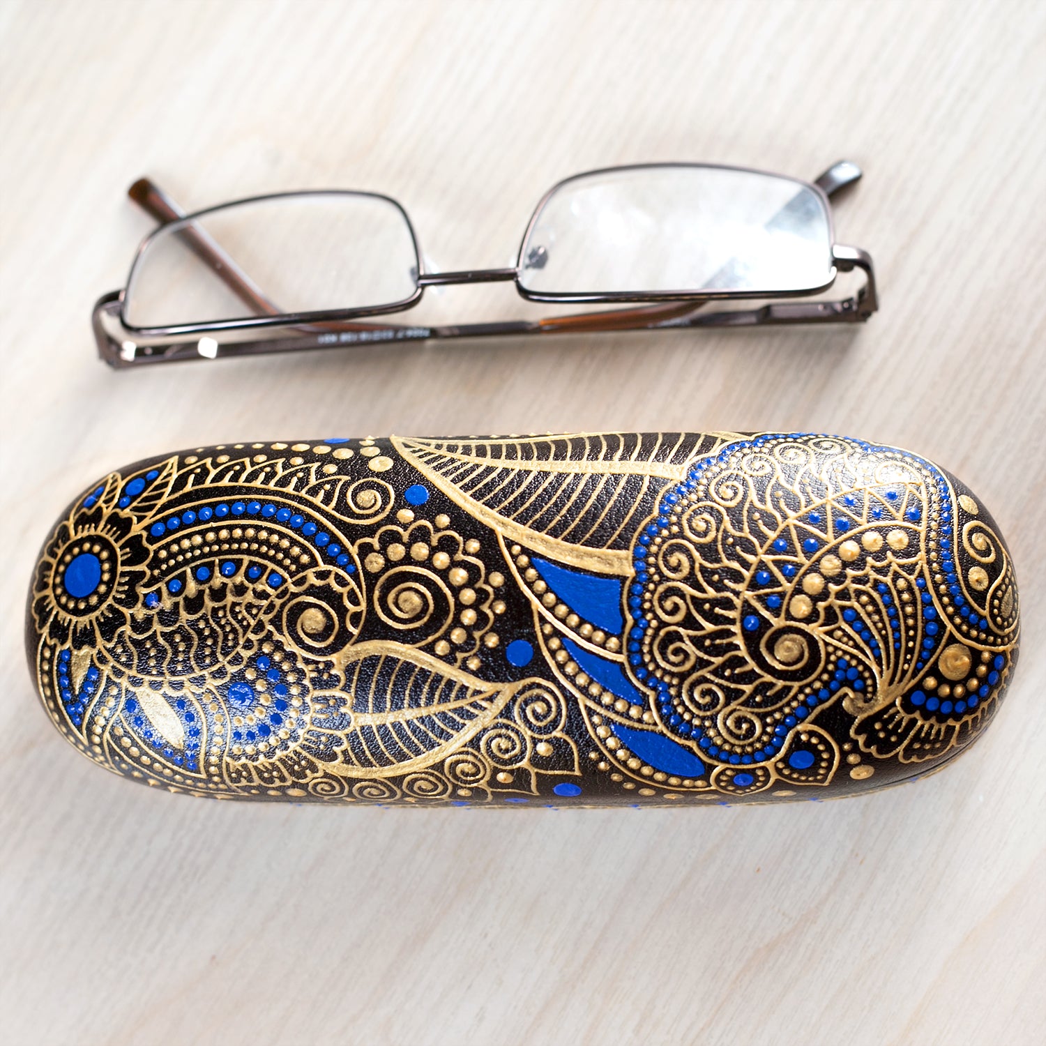 Hardshell case for glasses, hand-painted with blue and gold mehndi design. View from the top, reading glasses are on the side to show dimensions of the case.