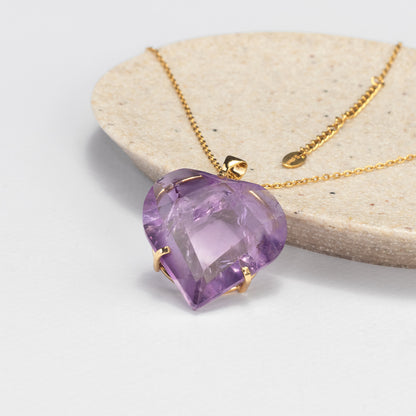 A heart-shaped purple amethyst pendant in a gold setting on a gold chain, displayed on a light beige stone slab against a white background.