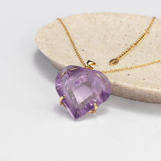 A heart-shaped purple amethyst pendant in a gold setting on a gold chain, displayed on a light beige stone slab against a white background.