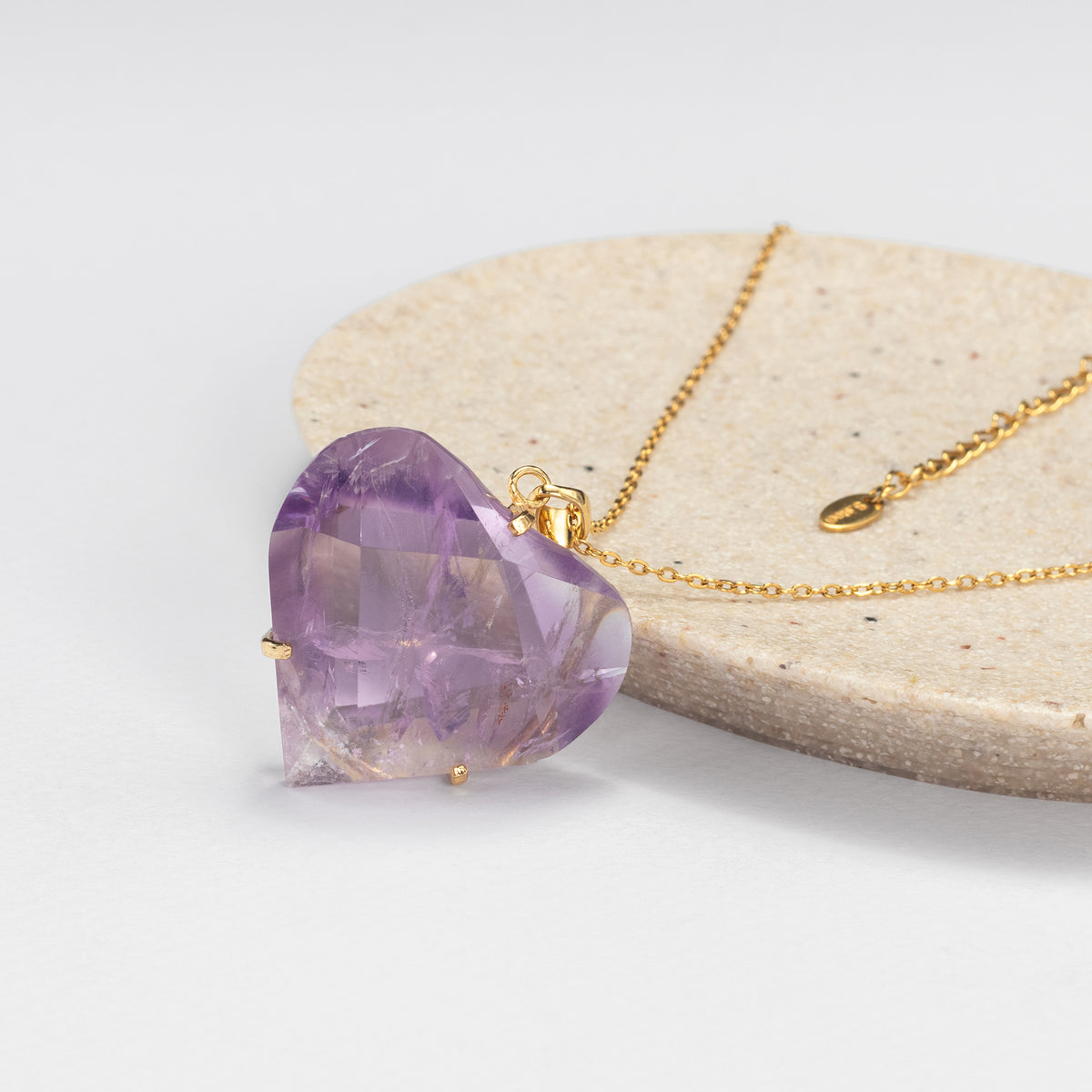 A faceted heart-shaped purple amethyst pendant on a delicate gold chain, displayed on a round beige stone slab against a clean white background.