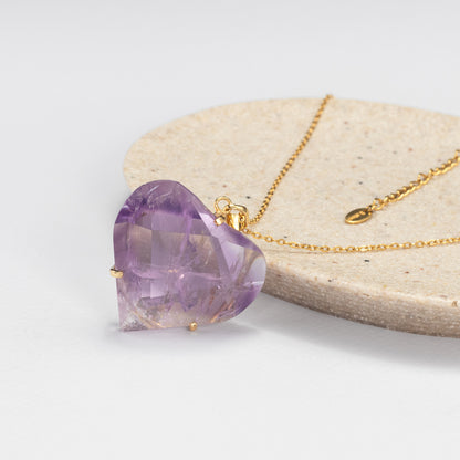 A faceted heart-shaped purple amethyst pendant on a delicate gold chain, displayed on a round beige stone slab against a clean white background.
