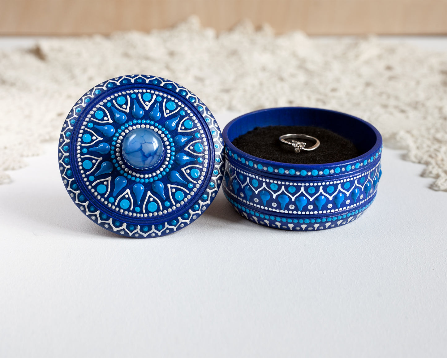 Wooden blue box with hand-painted design and blue agate stone on the lid. The box is open showing the ring inside. Standing on a white table