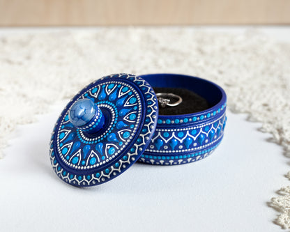 Wooden blue box with hand-painted design and blue agate stone on the lid. The box is open showing the ring inside. Standing on a white table