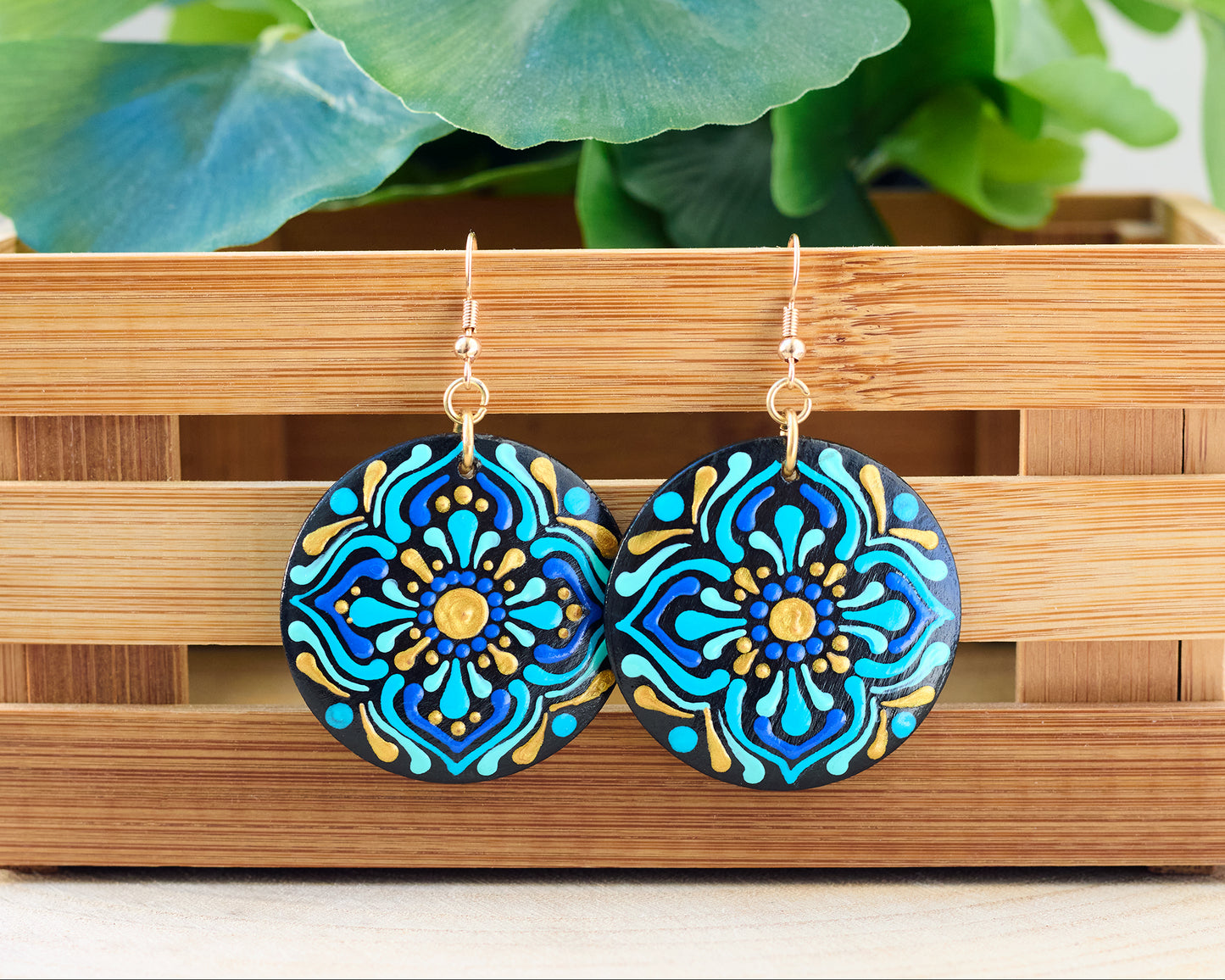Colorful mandala-patterned earrings on a wooden stand with green leaves in the background