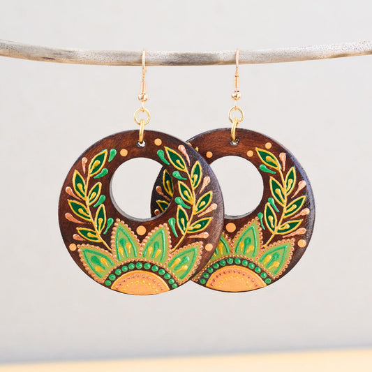Wooden earrings with green and gold leaf patterns hanging on a branch against a light gray background.