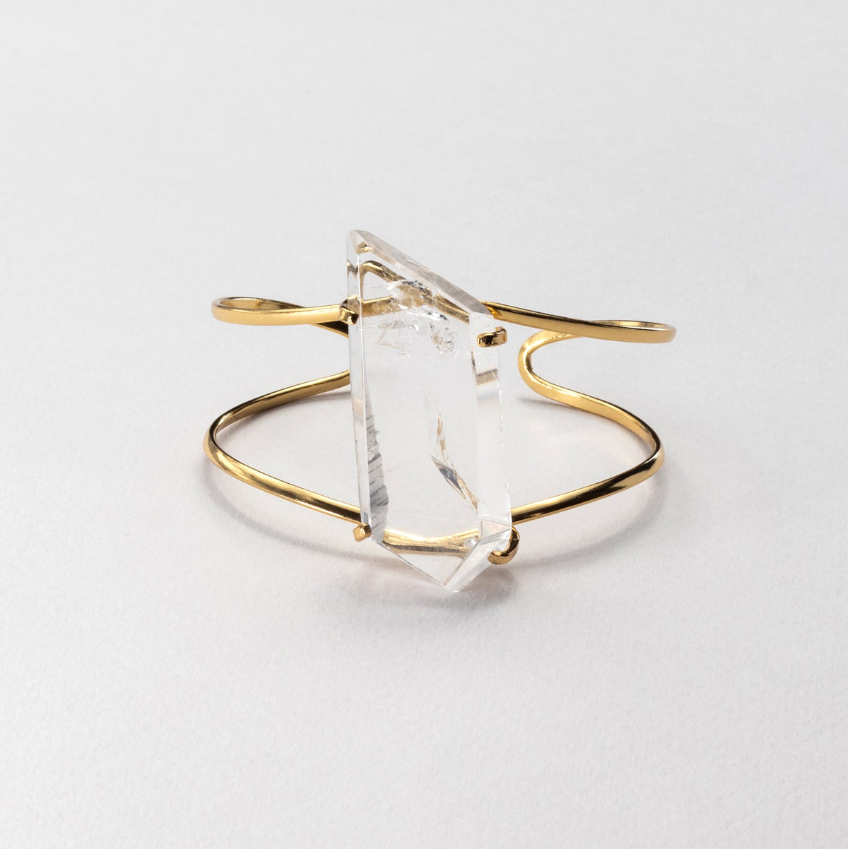 A gold-toned open cuff bracelet featuring a large, faceted clear crystal  on a clean, light gray background.