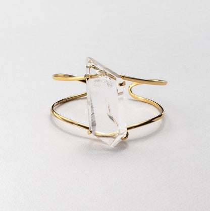 A gold-toned open cuff bracelet featuring a large, faceted clear crystal  on a clean, light gray background.