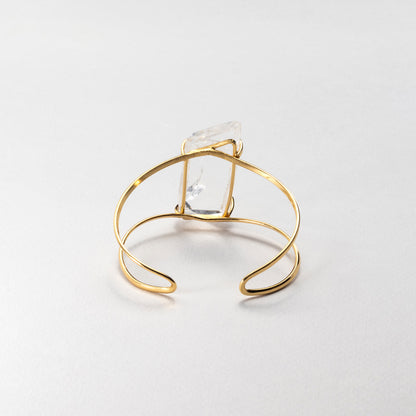 A gold wire wrap cuff bracelet featuring a clear, faceted rectangular crystal, photographed on a neutral light gray background.