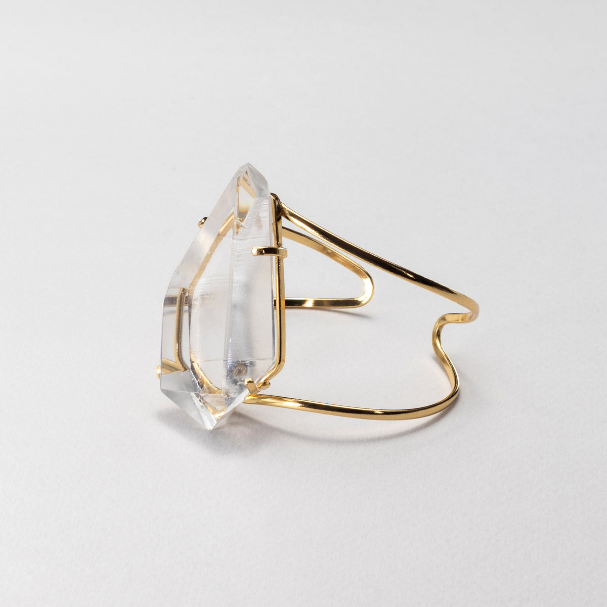 A gold cuff bracelet featuring a large, faceted clear crystal, photographed on a clean, light gray background.